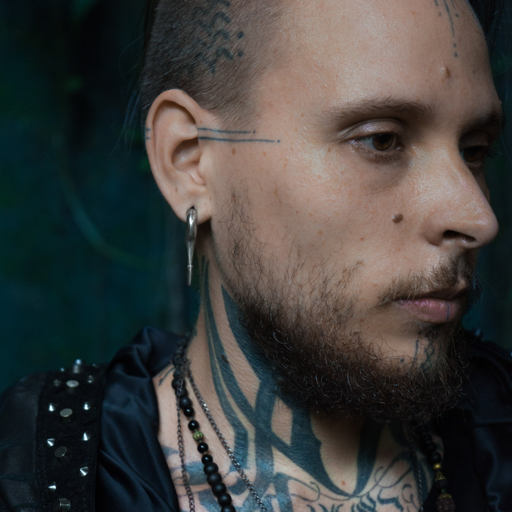 A person with tattoos on their face and neck is shown in profile. They have a beard, a pierced ear with a hoop earring, stretched ears adorned with CALYPSO Minimalist Keyhole Ear Weights in 925 Silver by Kaya Movement (2 gauge), and are wearing layered necklaces along with a studded garment. The background is a blurred, dark blue-toned setting.